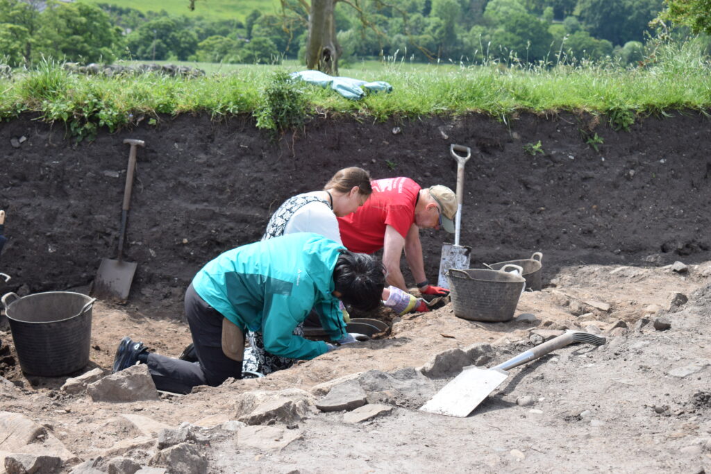 Three archaeological volunteers working in a ditch, surrounded  by spades and buckets. A grassy bank is in the background. 