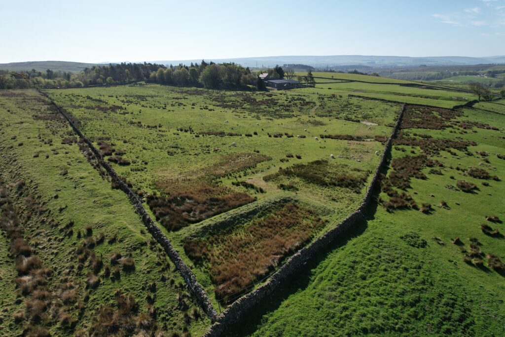 16:15Claude responded: Aerial view of a grassy upland landscape showing the rectangular earthwork outline of a Roman milecastle, with dry-stone field walls, grazing sheep, and a farm…Aerial view of a grassy upland landscape showing the rectangular earthwork outline of a Roman milecastle, with dry-stone field walls, grazing sheep, and a farmstead in the distance.