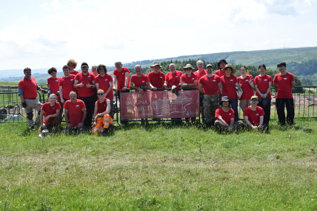 Group of archaeological volunteers all wearing red t-shirts, some are sitting in front of a green, metal fence and some are standing behind it. A banner that read 'The Magna Project' is attached to the fence and in the middle of the group of people. There is green grass in the foreground and rolling hills in the background.