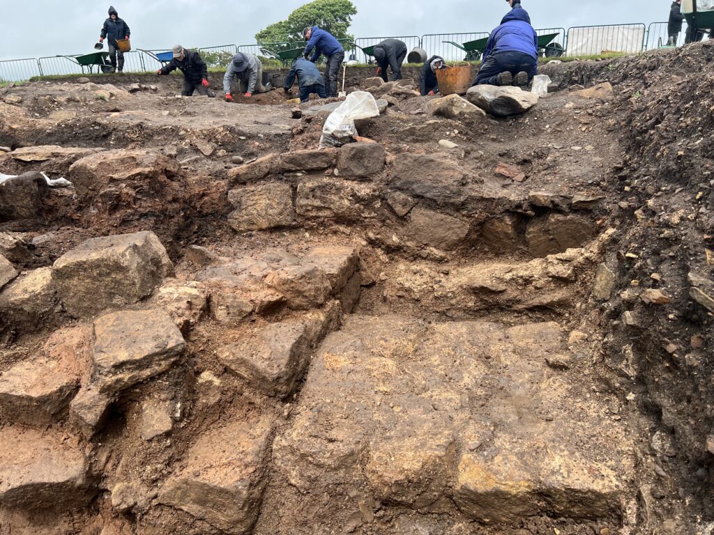 Close-up of multiple phases of stonework exposed in an excavation trench, with archaeologists digging and wheelbarrowing spoil in the background.