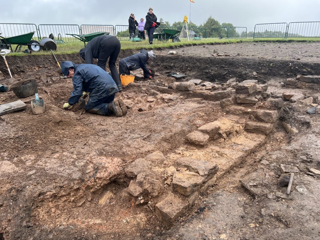Archaeologists kneel and crouch to excavate around exposed Roman stone wall foundations in a waterlogged trench on a rainy day, with onlookers watching from the edge above.
