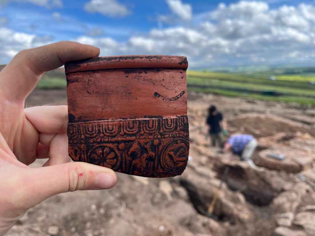 A close of up a sherd of samian ware, dark red and orange pottery from Gaul, stamped with a decorative pattern of flowers and birds. Archaeologist working in a stone filled trench in the background.