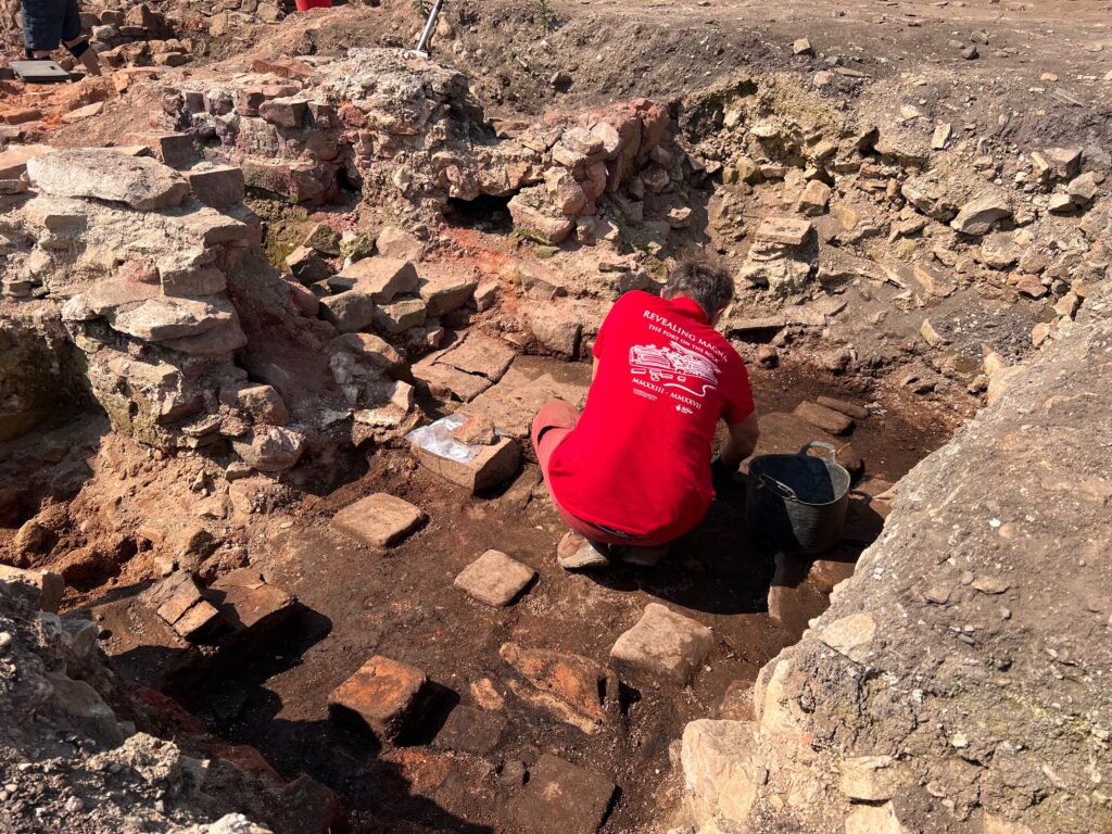 An excavator in a red "Revealing Magna" t-shirt kneels in a trench carefully uncovering a surface of Roman hypocaust system.