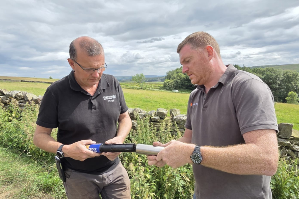 A Vindolanda Charitable Trust staff member and a colleague examine a piece of survey equipment outdoors, with dry-stone walls and open moorland behind them.