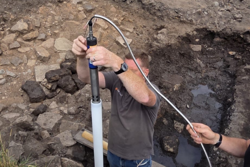 A person is fitting a long, metal tube connected to a long, silver wire into a white, plastic tube sitting in an archaeological trench. In the background is an open archaeological trench with rubble and soil visible.