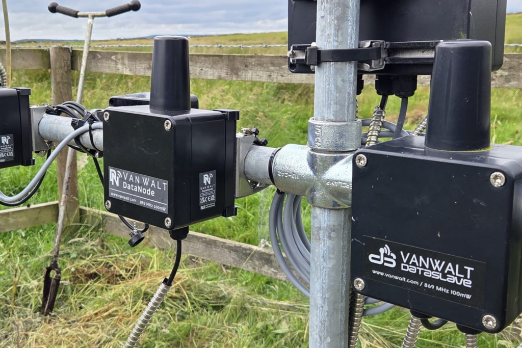 A close up of a weather station with silver poles and black data boxes with the words 'VanWalt Data Slave' written on. These are sitting in a grassy field.