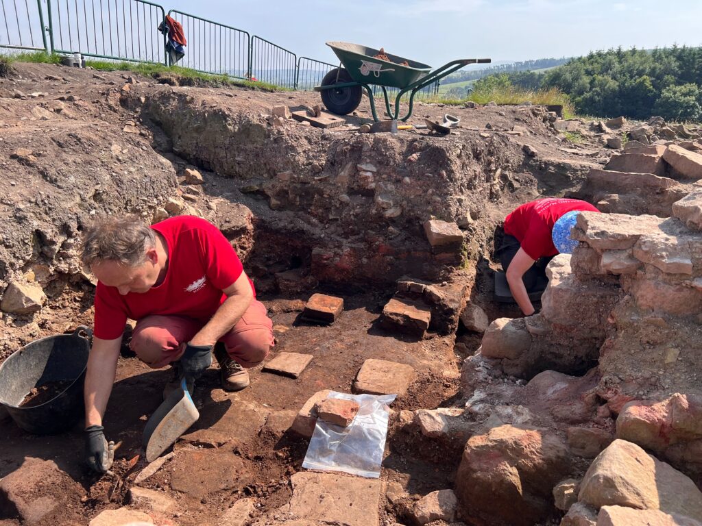 Two excavators in matching red t-shirts work in a sunlit trench exposing Roman hypocaust system, with a wheelbarrow and open hillside visible above.