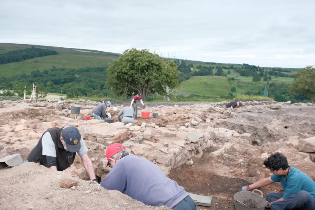 A team of archaeologists work across a large excavation site with exposed stone wall foundations, set against a green hillside landscape.