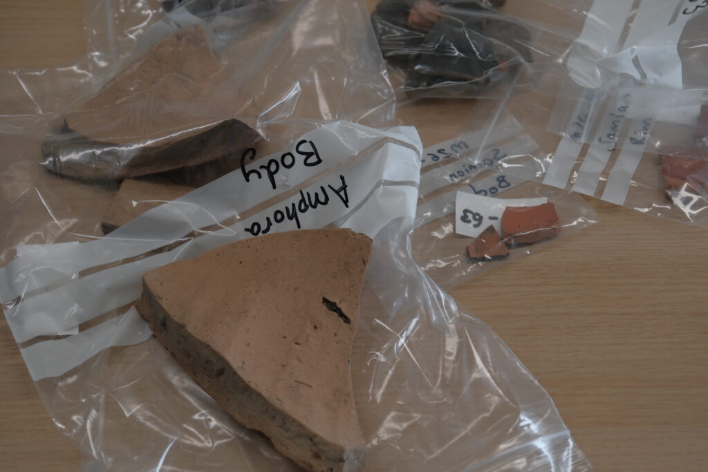 Labelled plastic bags containing pottery sherds, including an amphora body fragment and pieces of Samian ware, laid out on a table for examination.