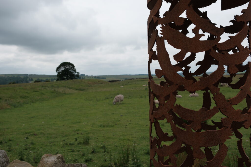 Sheep graze on open grassland viewed through a decorative rust-coloured laser-cut metal sculpture in the foreground.