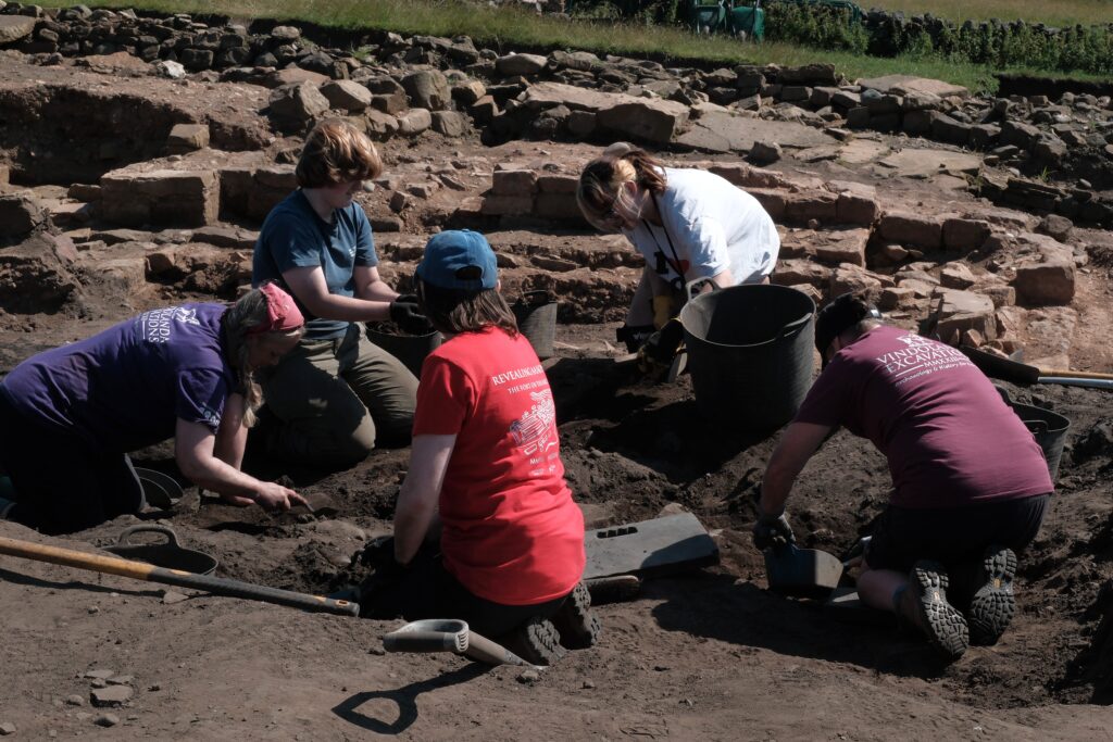 Five excavators kneel and crouch together in a sunlit trench, trowelling and filling buckets, with exposed Roman stonework visible behind them.
