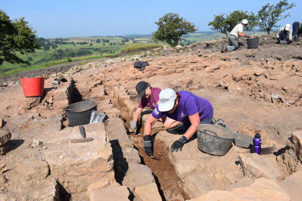 Two excavators trowel carefully between exposed stone wall foundations on a sunny hilltop site, with further team members working in the background and a sweeping green valley below.