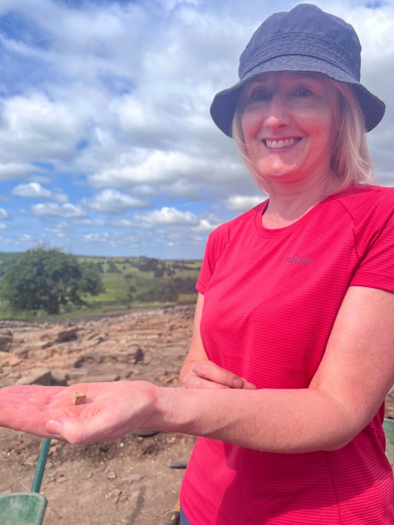 A smiling excavator wearing a red t-shirt and blue bucket hat holds out a small Roman die in her palm at the dig site, with the excavation trench and hillside visible behind her.