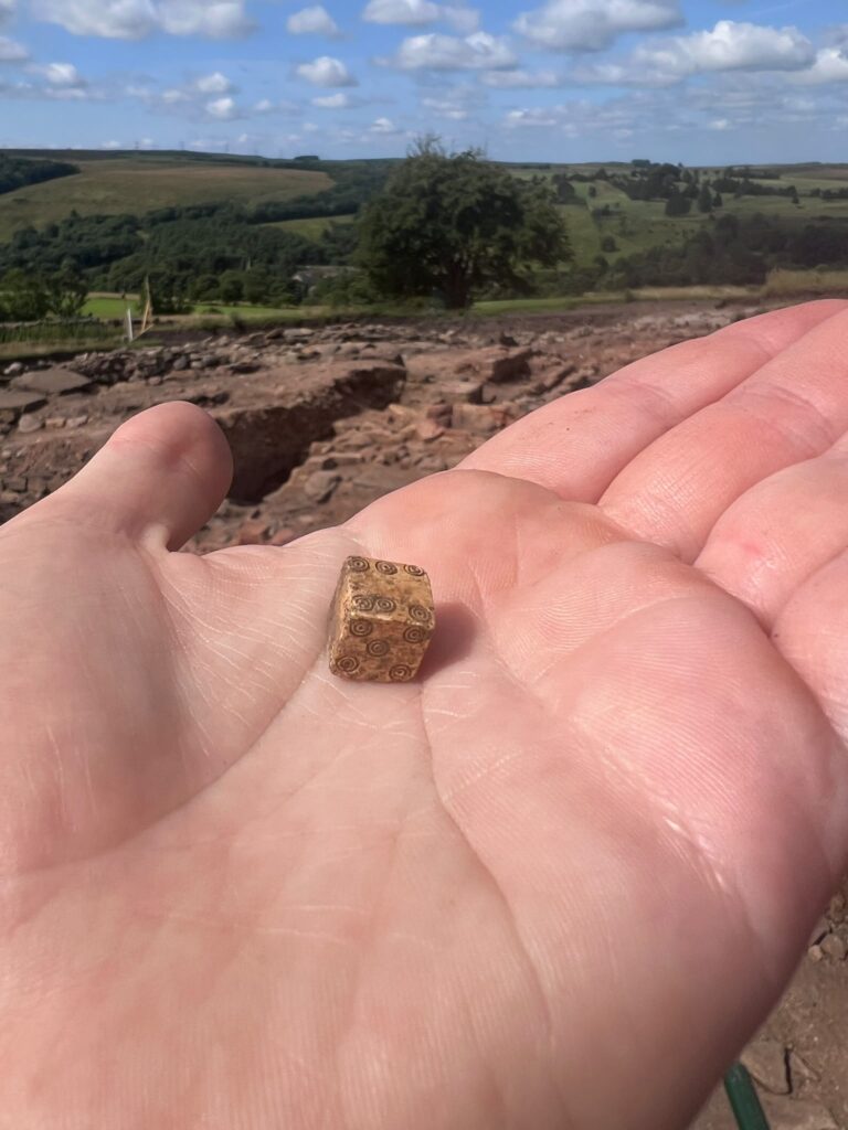 Close-up of a small Roman bone die decorated with concentric circle motifs on each face, resting in an open palm above the excavation site.