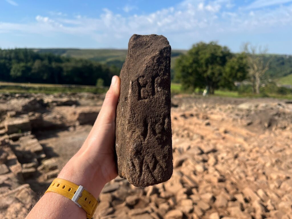 Left hand holding a brown stone with several ancient Roman letters inscribed on it in three rows.
