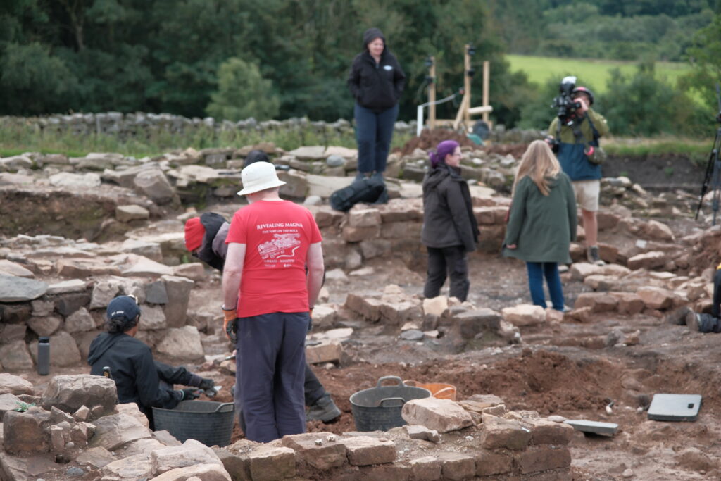 Person wearing a red 'Revealing Magna' t-shirt stands with their back to the camera. They are surrounded by archaeology, with a film crew in the background, filming an archaeologist and presenter. The rolling hills are in the background. 