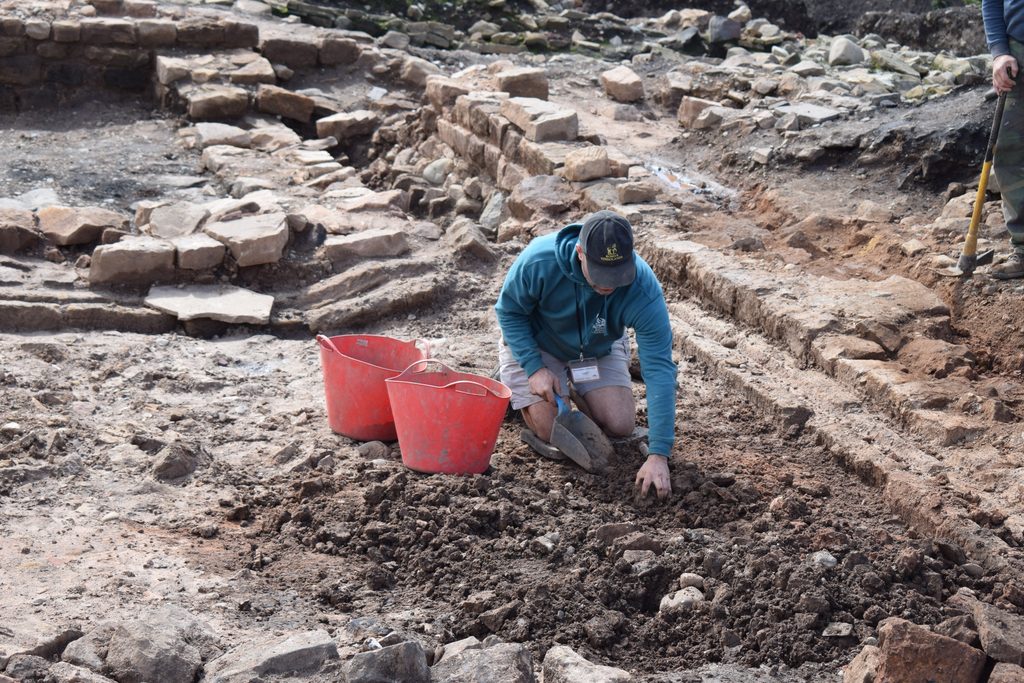 Person wearing a black cap, teal hoodie, and grey shorts kneels in an archaeological trench holding a scooper with two red buckets to the left of them. They are surrounded by archaeological remains, including a stone-cute drain running to the back and right of the image. 