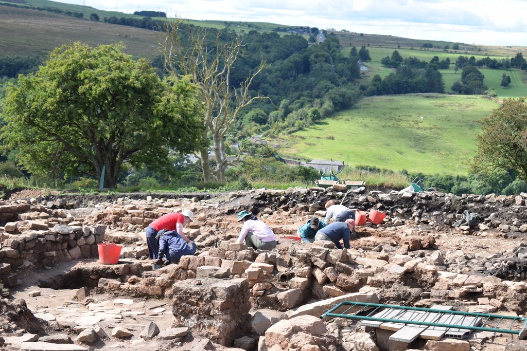 View across an archaeological excavation with stone wall remains. Volunteer archaeologists work in different sections of the trench, and there are trees and rolling hills in the background.