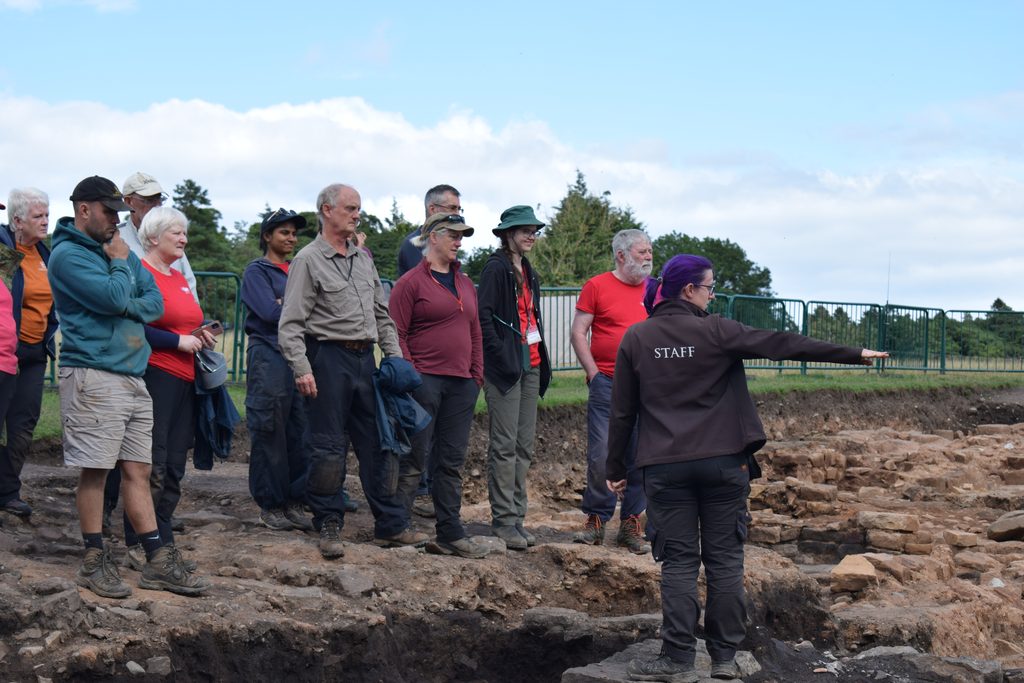 A person with purple hair wears a black jacket with 'staff' written on the back. They are stood in an archaeological trench pointing at a feature. Listening to them are volunteer excavators, wearing outdoor clothes. There is a green fence line and field in the background.