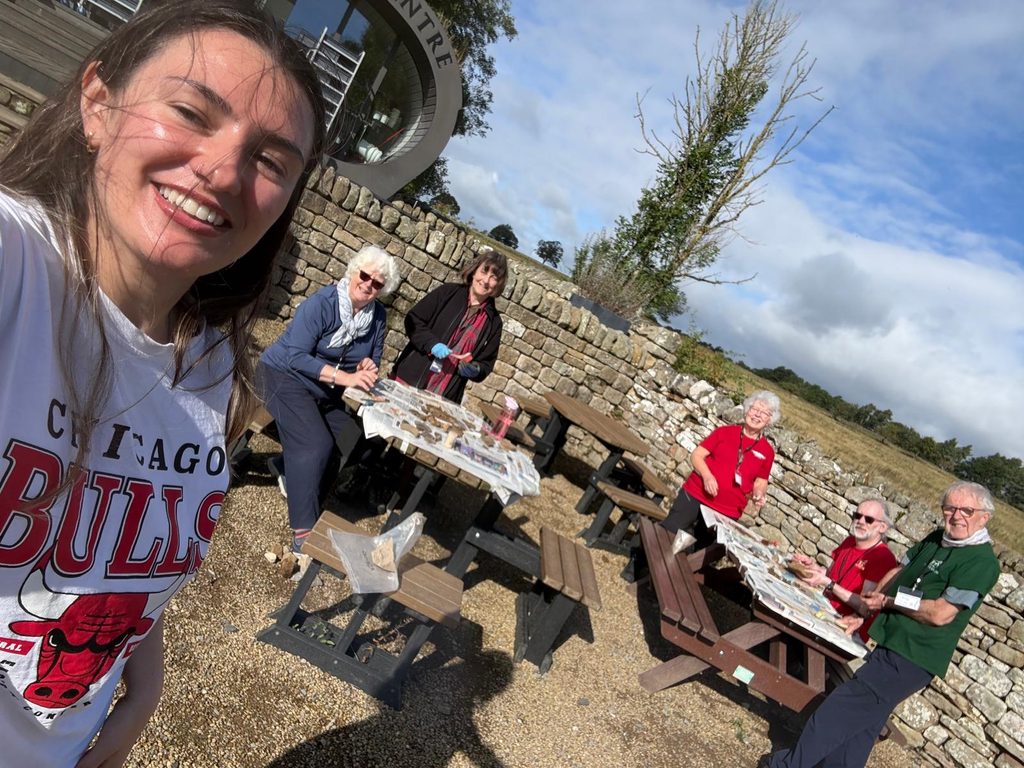 A selfie of a smiling staff member with a group of volunteers cleaning finds at picnic tables outdoors on a sunny day.