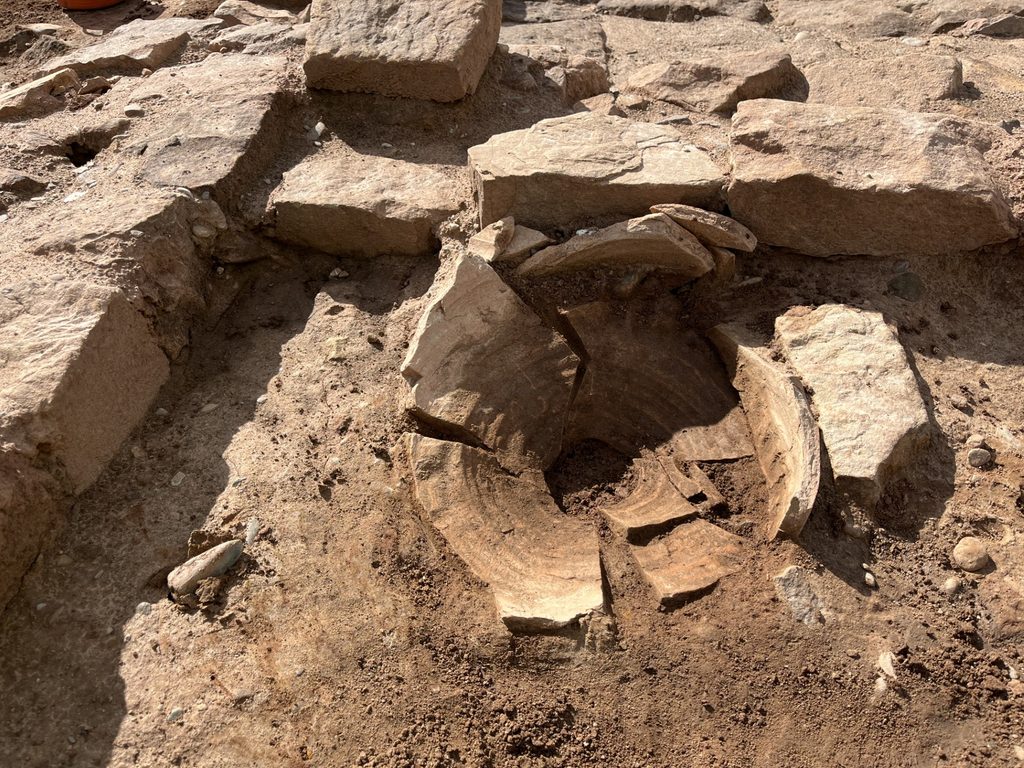A large pottery vessel found broken in situ within an excavation trench, its curved sherds still arranged in their original position among the stonework.
