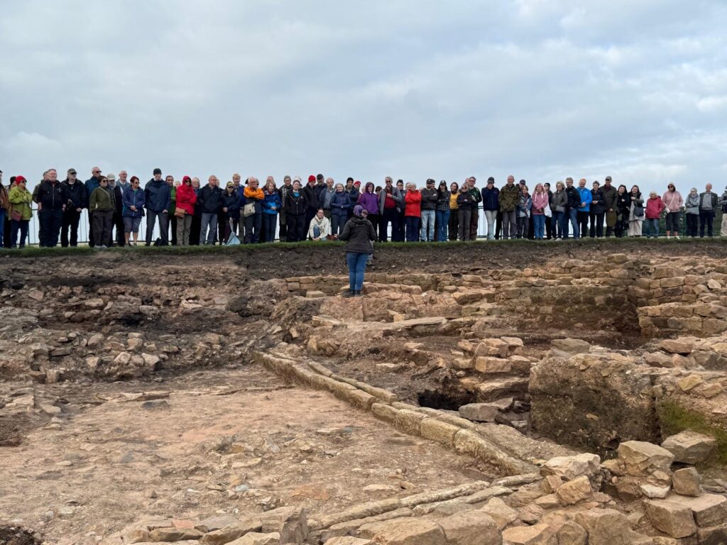 An archaeologist stands in a a large archaeological trench with exposed walls and drain features. Standing on the trench edge is a crowd of people listening to the archaeologist speak.
