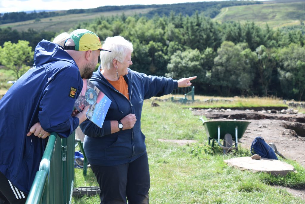A volunteer points out features across the excavation site to a visitor leaning on a barrier, with the open trench and hillside visible behind them.