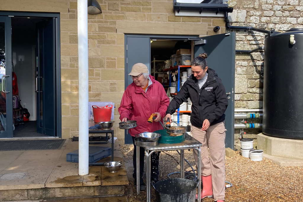A Vindolanda staff member and a volunteer wash finds using sieves and a hose at an outdoor finds-processing station beside the site building.