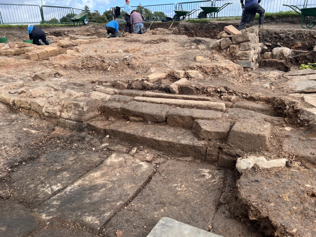 Close-up of neatly exposed Roman dressed stonework and flagstones in a large excavation trench, with team members working in the background.