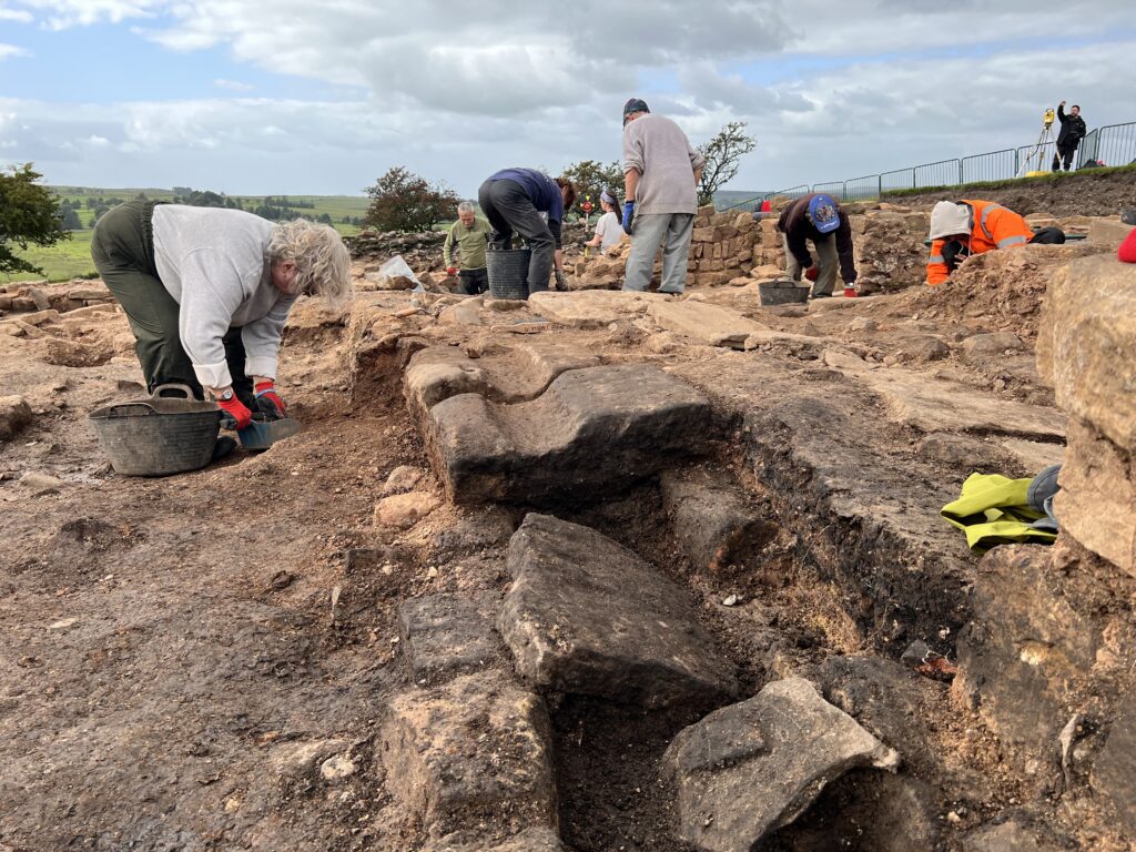 A wide team of excavators works across a large site uncovering substantial Roman stonework, with a surveyor using a total station on the perimeter and green hills beyond.