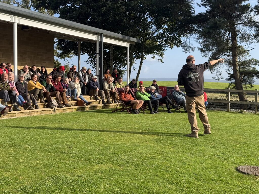 An image of a crowd sitting on some terraced steps in the setting sun facing a man in a black hoodie and brown trousers who is standing giving a talk. His back is to the camera and he is pointing to the right.