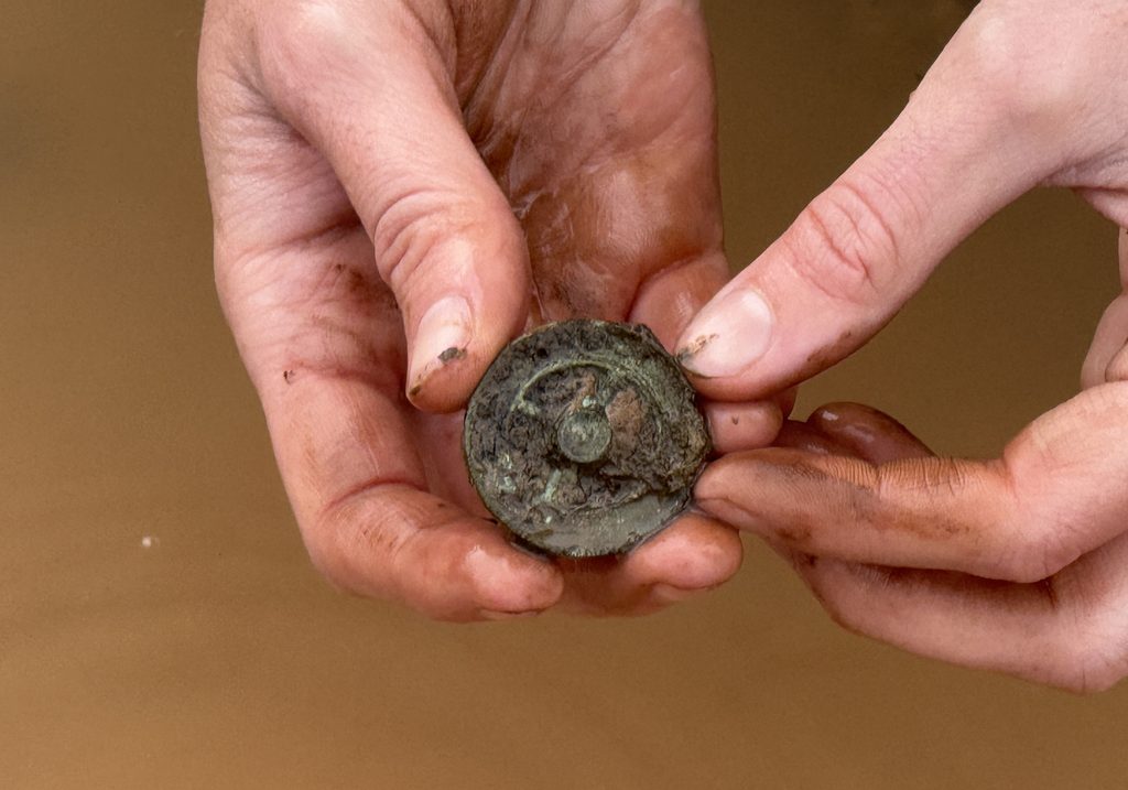 Two muddy hands hold a circular Roman copper-alloy wheel brooch, freshly retrieved from the ground, its spoked design still visible through the soil.