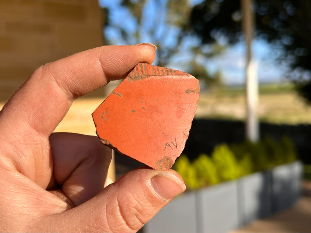 Close up of a sherd of Roman pottery being held between a finger and thumb. The piece of pottery is a rough square shape, a deep orange colour, with the letters 'AV' graffitied in the right bottom corner.